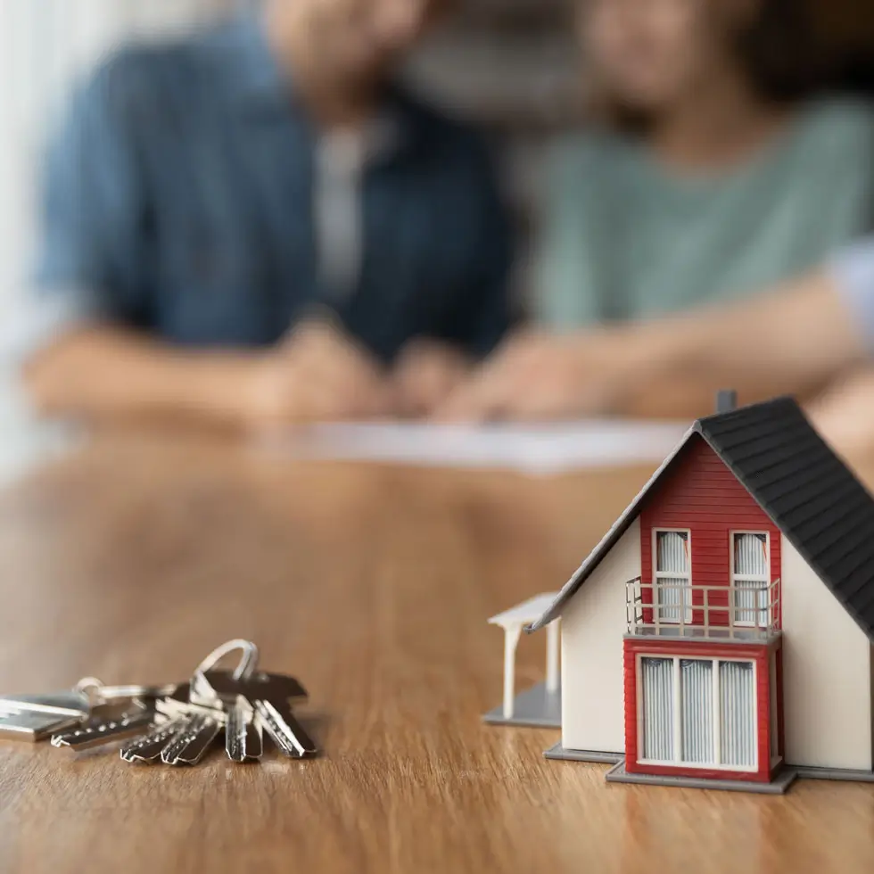 Nahaufnahme eines Schlüssels und eines winzigen Spielzeughauses auf einem Tisch. Im Hintergrund: Ehepaar bei der Beratung für den Kauf eines Hauses mit einem Immobilienmakler